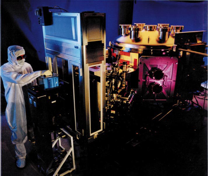 A technician in a cleanroom bunny suit operating an extreme ultraviolet lithography tool with a gold-colored vacuum chamber